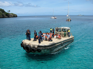 Asylum seekers arrive at Christmas Island on a small flat boat, wearing life jackets, kneeling in a line and being escorted by what appear to be immigration staff.