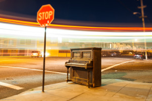 A piano on a street corner at night, next to a stop sign, with blurred, out of focus car lights passing by