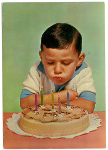An old photo, probably from the 60s, of a nerdy looking boy blowing out the candles on a birthday cake