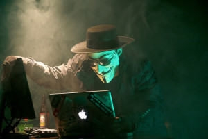 A stereotypical security hacker, using a computer in a dark room while wearing a guy flakes mask and a black hat.