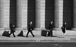 Four people in suits walking in front of a building with tall, thick columns, possibly a bank or museum, with long shadows in the early morning light.
