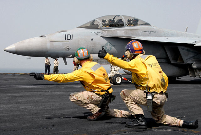 A fighter jet on an aircraft carrier being signalled to launch by two ground staff. Launching a fighter jet is a costly deployment, just as deploying a monolith may be.